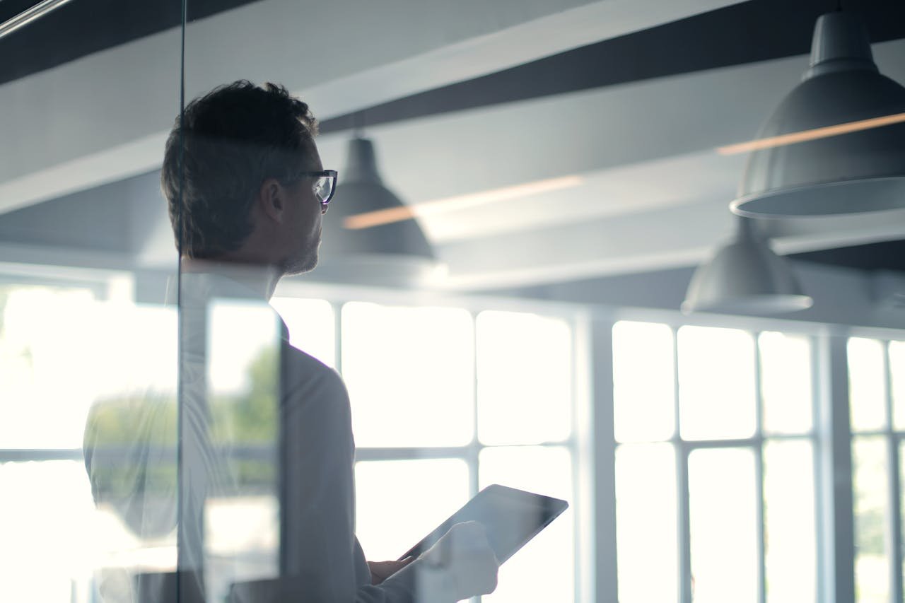 about-02 A businessman holding a tablet in an office, looking thoughtfully through a glass wall.